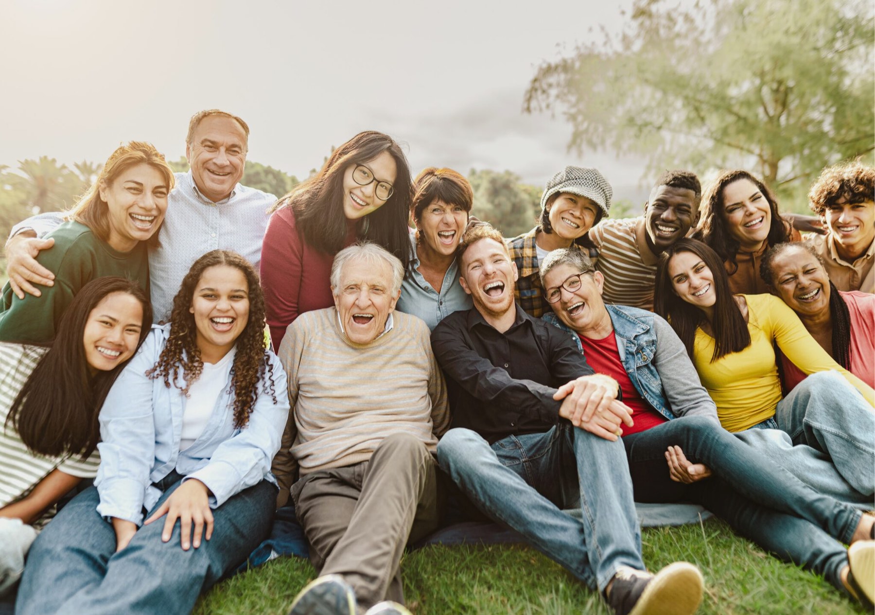 A large group of people of different ages and backgrounds sitting together on the grass in a park, laughing